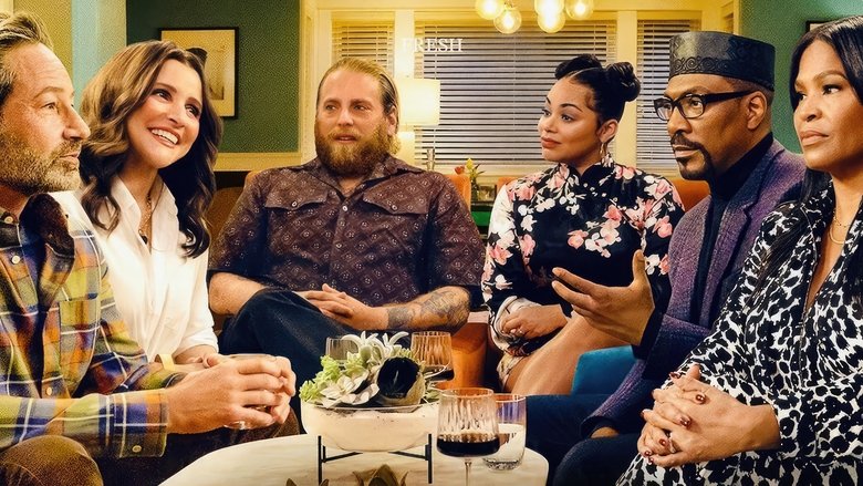 A couple sits on a couch, surrounded by family, tension thick in the air