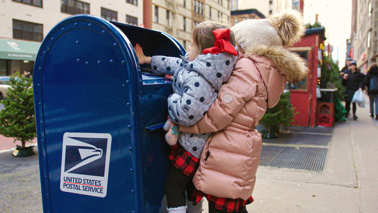 A volunteer in a festive sweater sorts through stacks of handwritten letters to Santa, their faces lit by the warm glow of holiday lights.