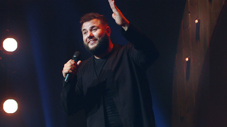 Mo Amer on stage, mid-performance, gesturing with his hands as he tells a story