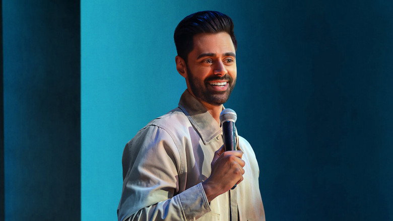 Hasan Minhaj mid-performance, arms outstretched, bathed in stage light