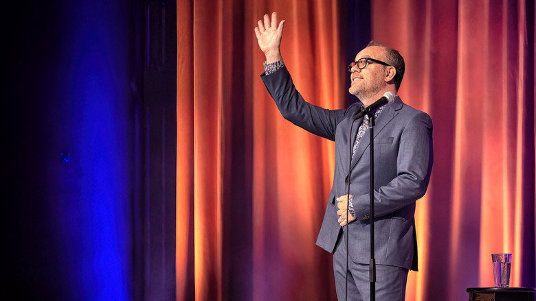 Tom Papa on stage, mid-gesture, bathed in warm light, the audience blurred in the background