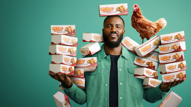 Mo Gilligan sits in a fast-food restaurant booth, holding a piece of fried chicken, his expression a mix of amusement and unease.