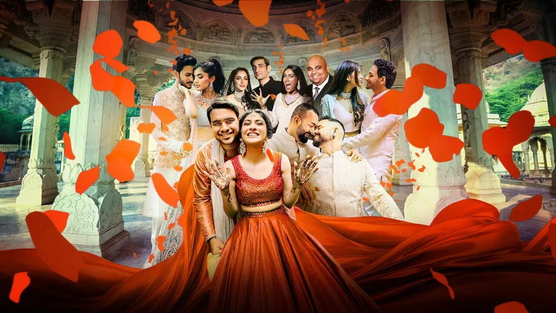 A bride in a lavish red and gold lehenga stands before a grand floral arch, her hands clasped in front of her, surrounded by attendants in matching outfits