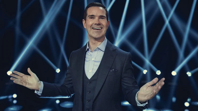 A dimly lit stage, Carr standing with hands in pockets, his face half in shadow, the audience barely visible beyond the spotlight.