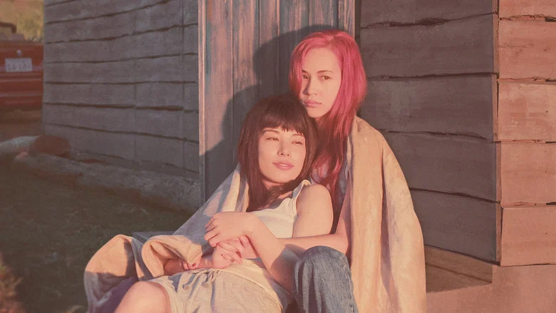 Two women sit in a dimly lit room, the glow of a television casting blue light across their faces. The tension between them is palpable, a mix of fear and something unspoken.