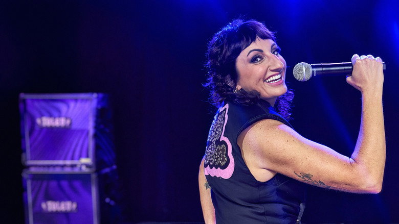 Steph Tolev on stage in a black pleather jumpsuit, mid-laugh, arms outstretched as if inviting the audience into her filth