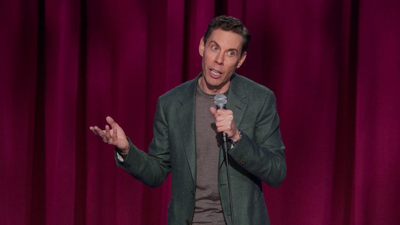 Ryan Hamilton on stage, mid-laugh, gesturing with his hands as if explaining the exact moment the bus made contact.