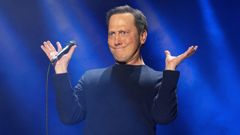 Rob Schneider on stage, mid-performance, gesturing with his hands as he tells a joke