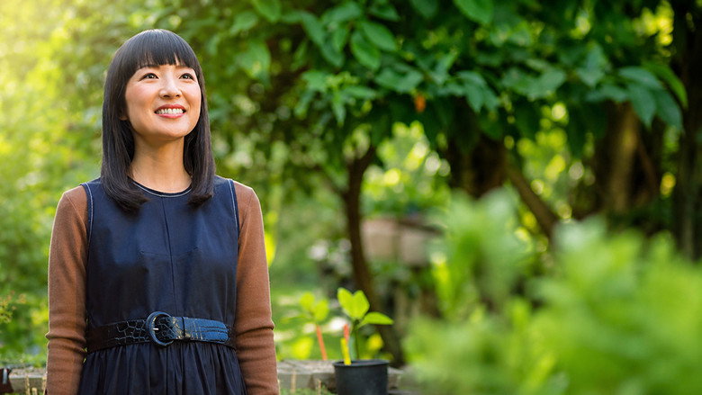 Marie Kondo kneels on the floor of a cluttered living room, her hands resting on a pile of fabric as she speaks softly to a family seated across from her.