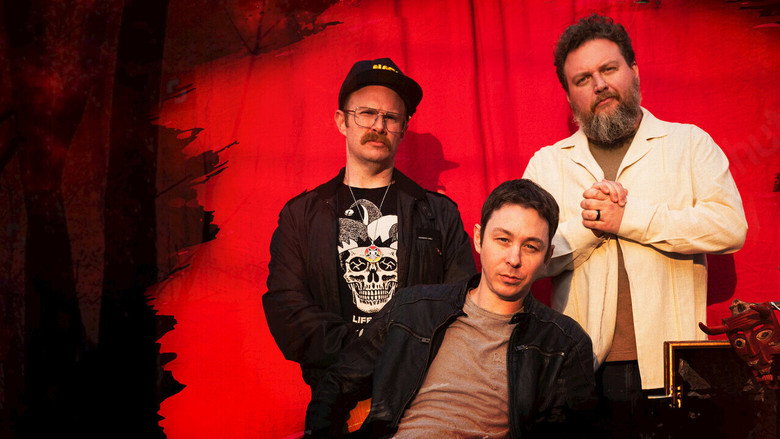 The hosts sit in a dimly lit studio, their faces half in shadow, as if caught between a punchline and a scream.
