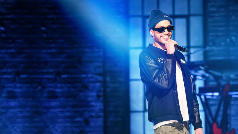 Pete Davidson on stage, mid-joke, with a microphone in hand, the audience blurred in the background