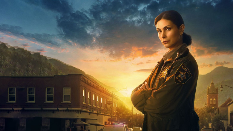 Mickey Fox sitting at a cluttered, dimly lit wooden kitchen table, looking tired, with her father blurred in the background near a window.