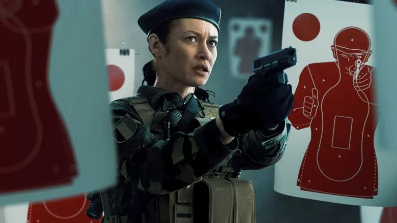 A soldier in uniform stands in a dimly lit room, her face half-shadowed, staring intently at something off-camera.