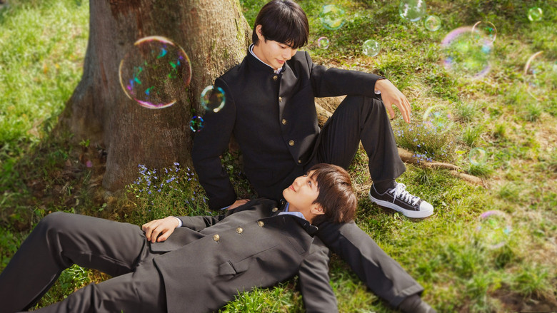 Two boys sit side by side on a bench, their bicycles leaning against it, as one stares intently at the other, who looks away with a faint, conflicted smile.