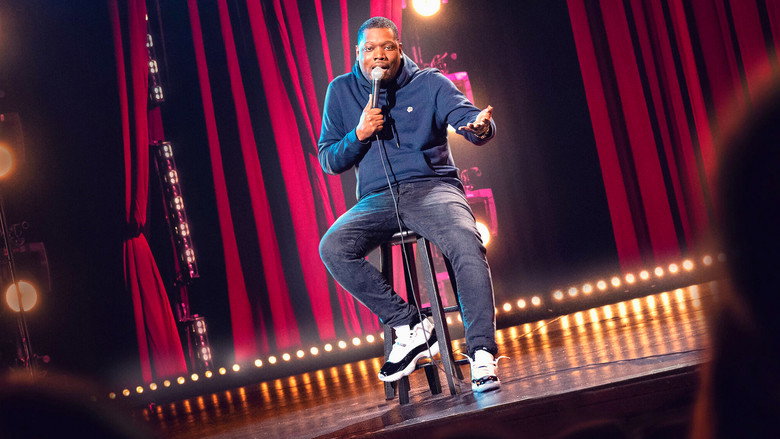 Michael Che on stage, mid-performance, bathed in warm light, gesturing with one hand while the other rests on his chest.
