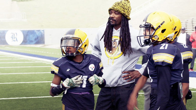 Snoop Dogg in a huddle with young football players, his face serious under a backward cap