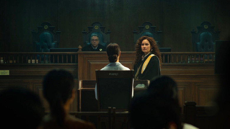 A dimly lit courtroom, the camera angled low to emphasize the looming presence of the judge’s bench, while Jittri stands in the foreground, her back to the camera, her posture radiating controlled menace.