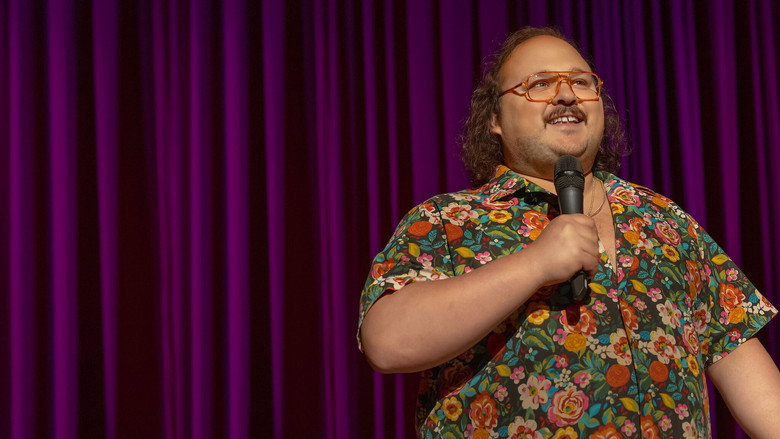 A dimly lit stage, the comedian mid-gesture, his face half in shadow, the audience a blur of indistinct shapes.
