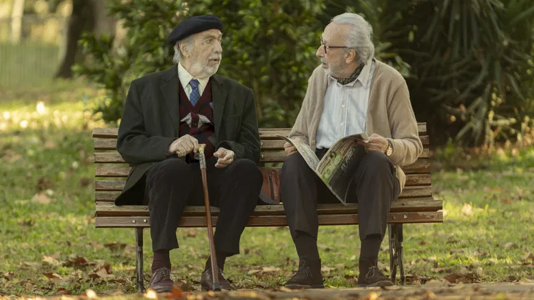 Two elderly men sit on a park bench, their faces half-lit by dappled sunlight, one gesturing emphatically while the other listens with a mix of amusement and exhaustion.