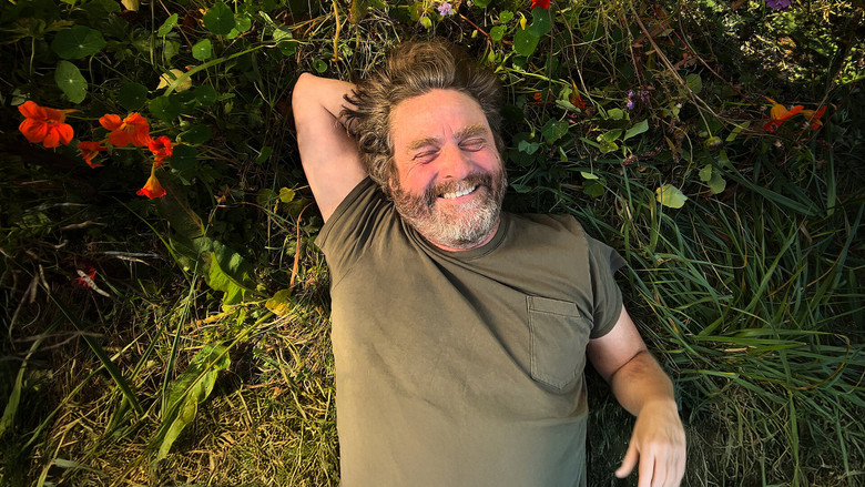 Zach Galifianakis kneels in a garden bed, surrounded by children, all of them grinning at something just out of frame