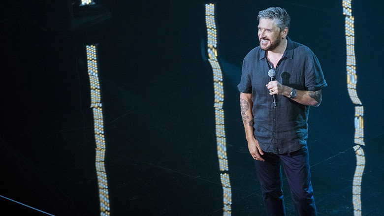 Craig Ferguson on stage, mid-gesture, in a dimly lit room with a stool and microphone