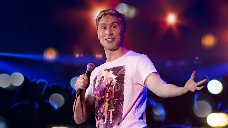 Russell Howard on stage, mid-gesture, bathed in warm light, the audience a blur of faces beyond the spotlight.