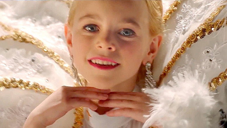 A young girl in a sparkly pageant dress stands under a spotlight, her face half in shadow