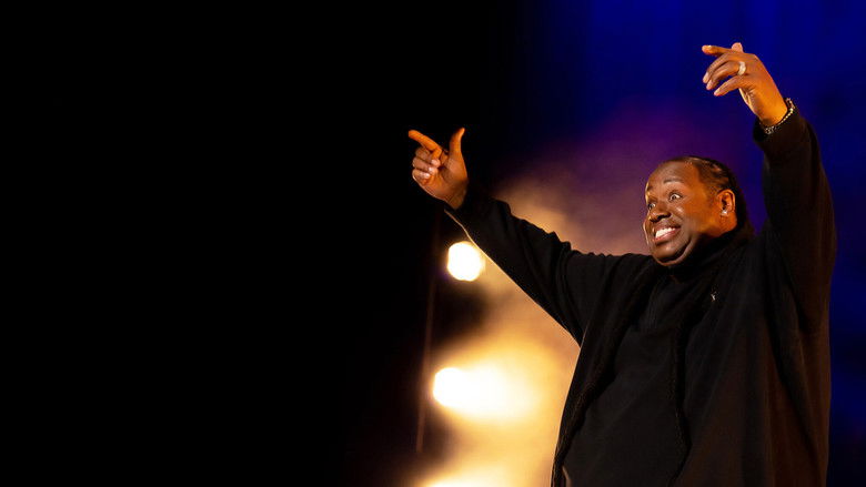 Bruce Bruce on stage, leaning back on a stool with a microphone in hand, mid-laugh