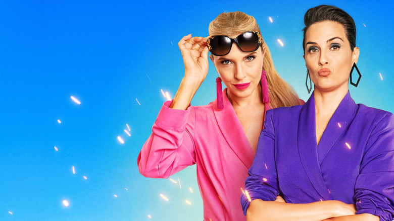 Two women sit in a circle of chairs, one mid-eye-roll, the other gripping her knees, under harsh fluorescent lighting