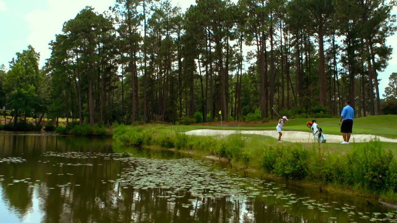 A seven-year-old golfer in a red cap crouches over a putt, his shadow stretching long across the green.