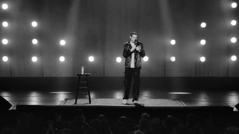Pete Davidson on stage in black and white, mid-gesture, the spotlight casting long shadows across his face