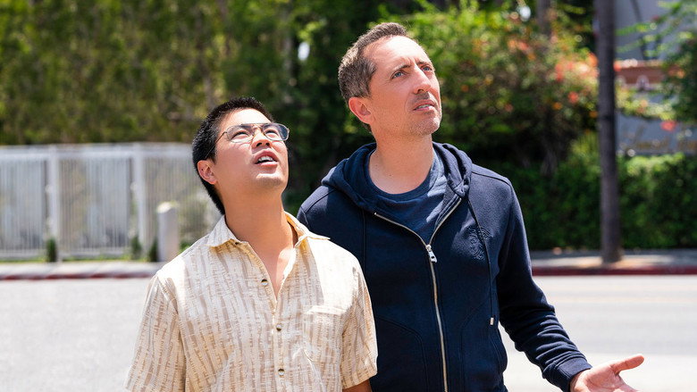Gad Elmaleh standing in a sunlit Los Angeles backyard, looking slightly out of place in a polo shirt and jeans, while a teenager lounges in the background.