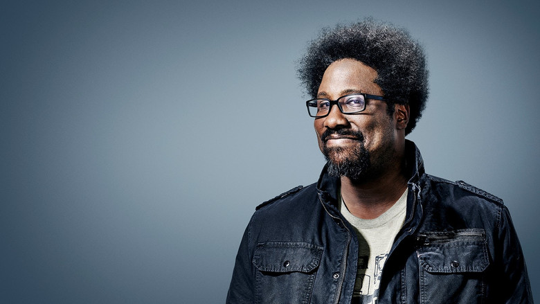 W. Kamau Bell on stage, holding up a children's book with a wry expression