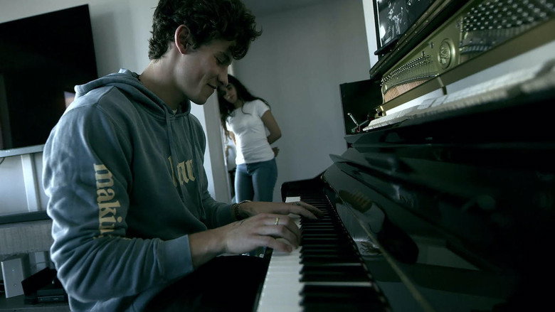 Shawn Mendes sitting alone in a dimly lit hotel room, his face half in shadow, staring at his phone.