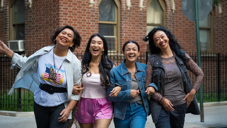 The four women sit around a cluttered kitchen table, laughing over takeout containers, their faces lit by the warm glow of a single overhead bulb.