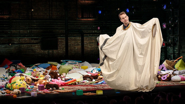 Birbiglia on stage, mid-story, gesturing toward the couch that becomes a central metaphor