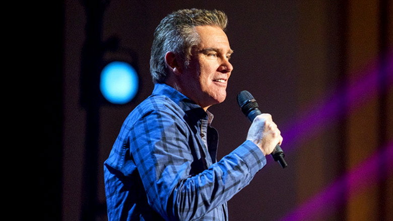 Brian Regan on stage, mid-joke, hands gesturing as if explaining something absurd