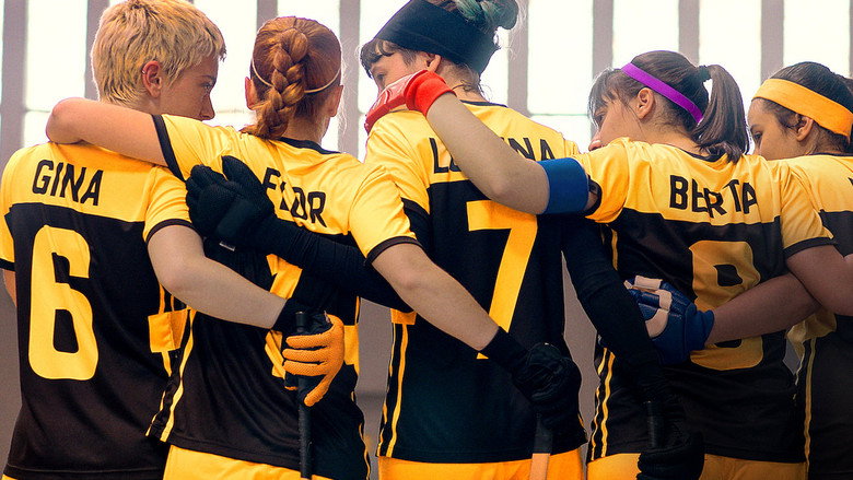 A group of girls in roller hockey gear stand in a huddle, their faces determined, the rink stretching out behind them in the golden light of late afternoon.