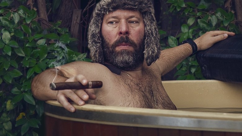Bert Kreischer and a guest sitting on a couch in a dimly lit cabin, mid-conversation