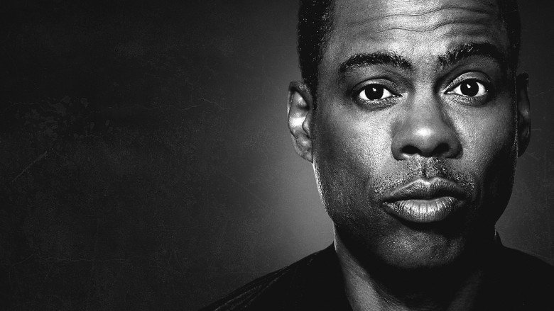 A lone spotlight illuminates Chris Rock on stage, his expression caught between defiance and exhaustion.
