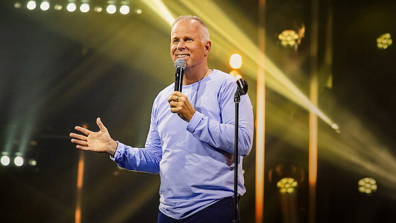 Gerry Dee standing on stage under warm blue lighting, holding a microphone