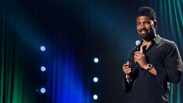 Deon Cole mid-laugh on stage, the audience blurred in the background