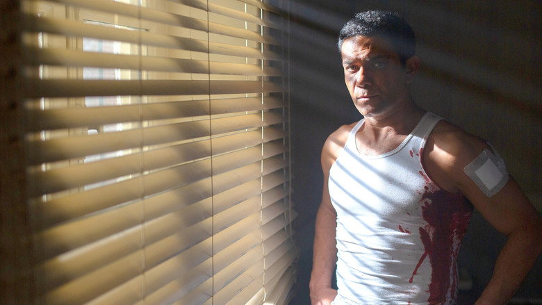 A dimly lit room where Yehia stands over a table covered in photographs, his face half in shadow, the weight of the past pressing down on him.
