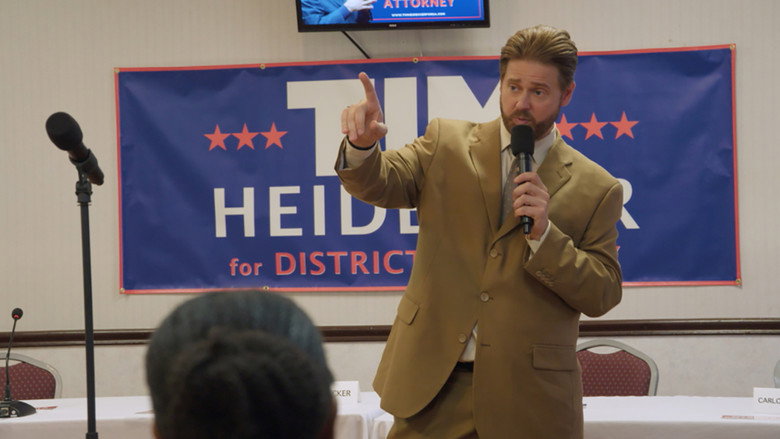 Heidecker stands at a podium, his face half-lit by a cheap campaign banner, the words "Mister America" stretched awkwardly across it.