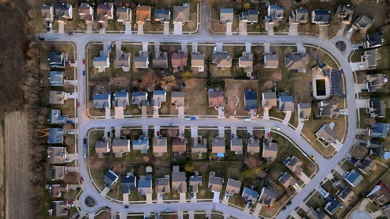A dimly lit suburban street at night, the glow of a porch light casting long shadows across a driveway where a lone figure stands, their face obscured.