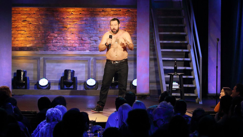 Bert Kreischer mid-laugh, arms wide, shirtless under stage lights