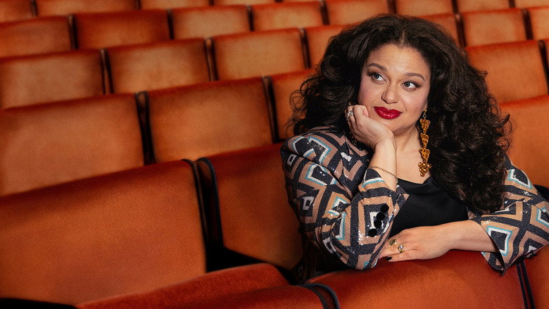 Michelle Buteau on stage at Radio City Music Hall, mid-laugh, arms outstretched as the audience glows in the background