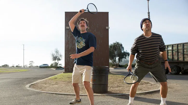Two men stand in a dimly lit parking lot, one holding a makeshift paddle, the other watching with a mix of amusement and something harder to name