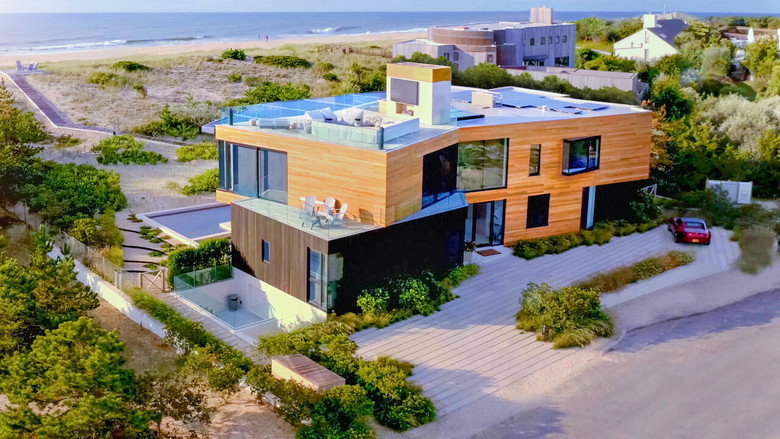 A sweeping aerial shot of a modern Hamptons mansion, its glass walls reflecting the golden light of a summer sunset.