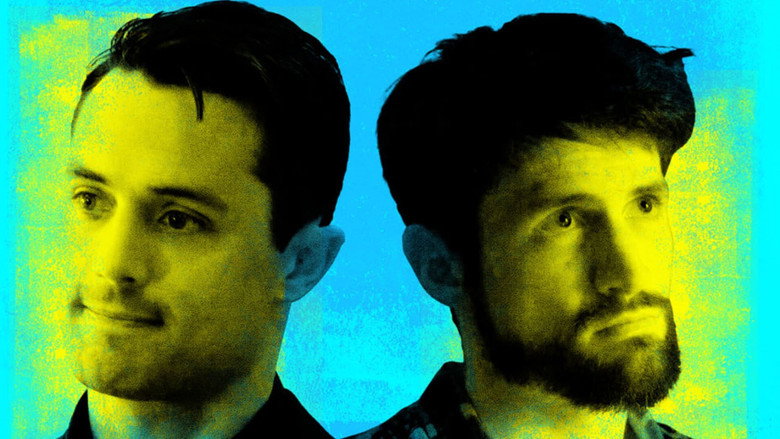 Two men sit on a couch, one mid-laugh, the other staring blankly ahead, the glow of a television screen casting long shadows across their faces.